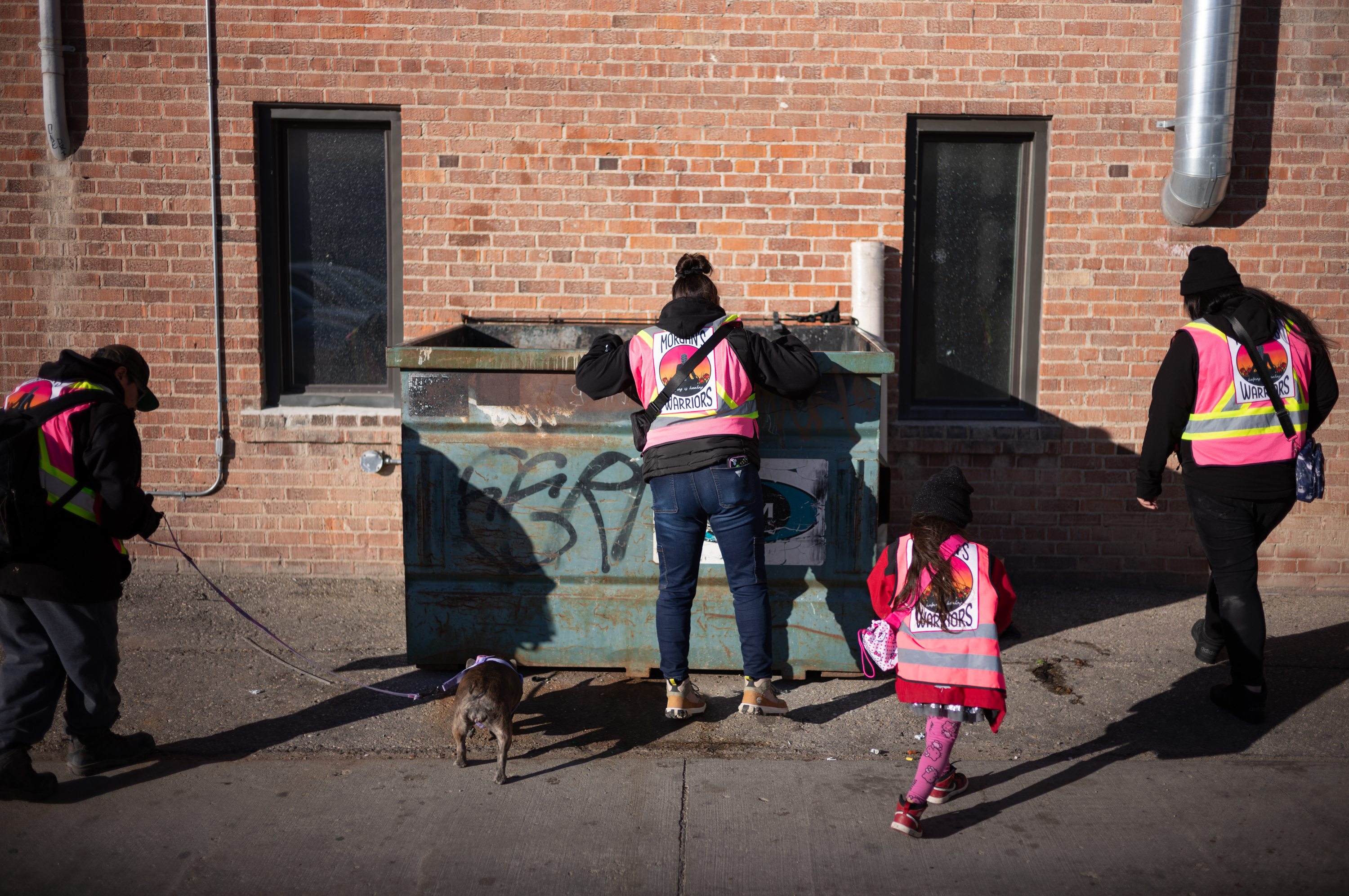 Women and girls in pink fluorescent jackets search through a large rubbish bin