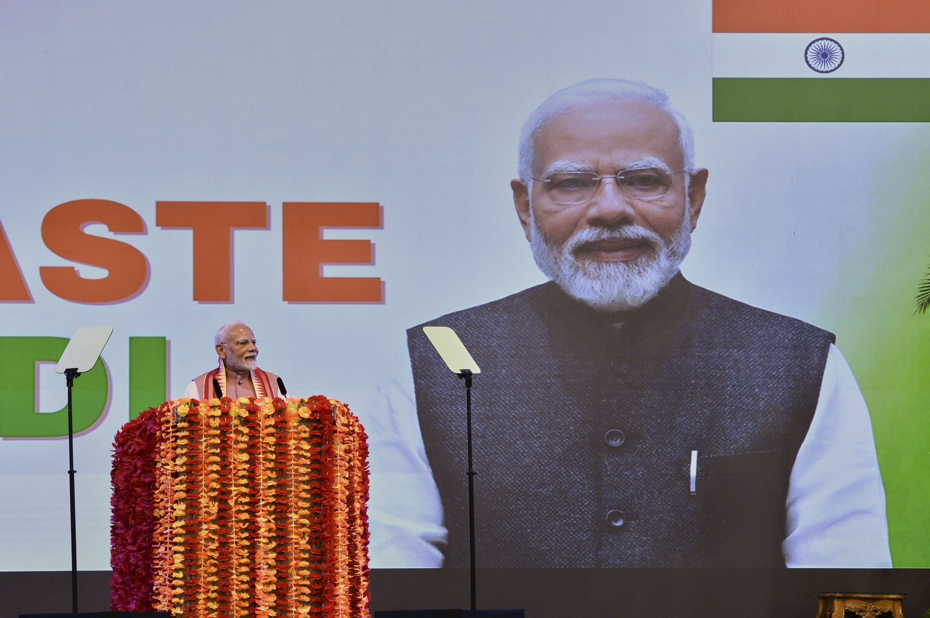 Narendra Modi speaks at a podium. Behind him is a screen projecting his picture and an Indian flag.