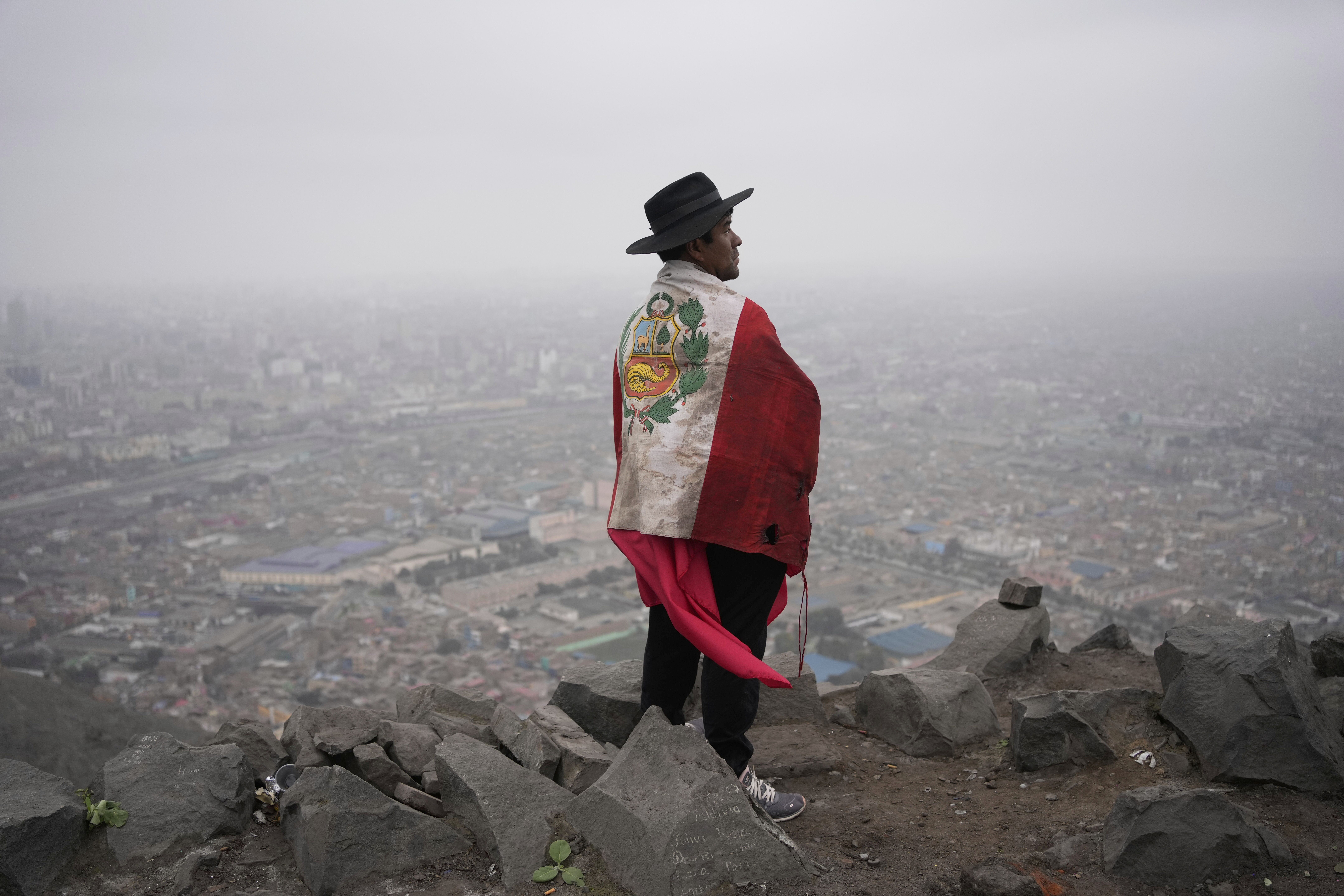 A Peruvian man wearing a broad black hat, with a Peruvian flag draped over his shoulders, stands on a hill overlooking Lima.