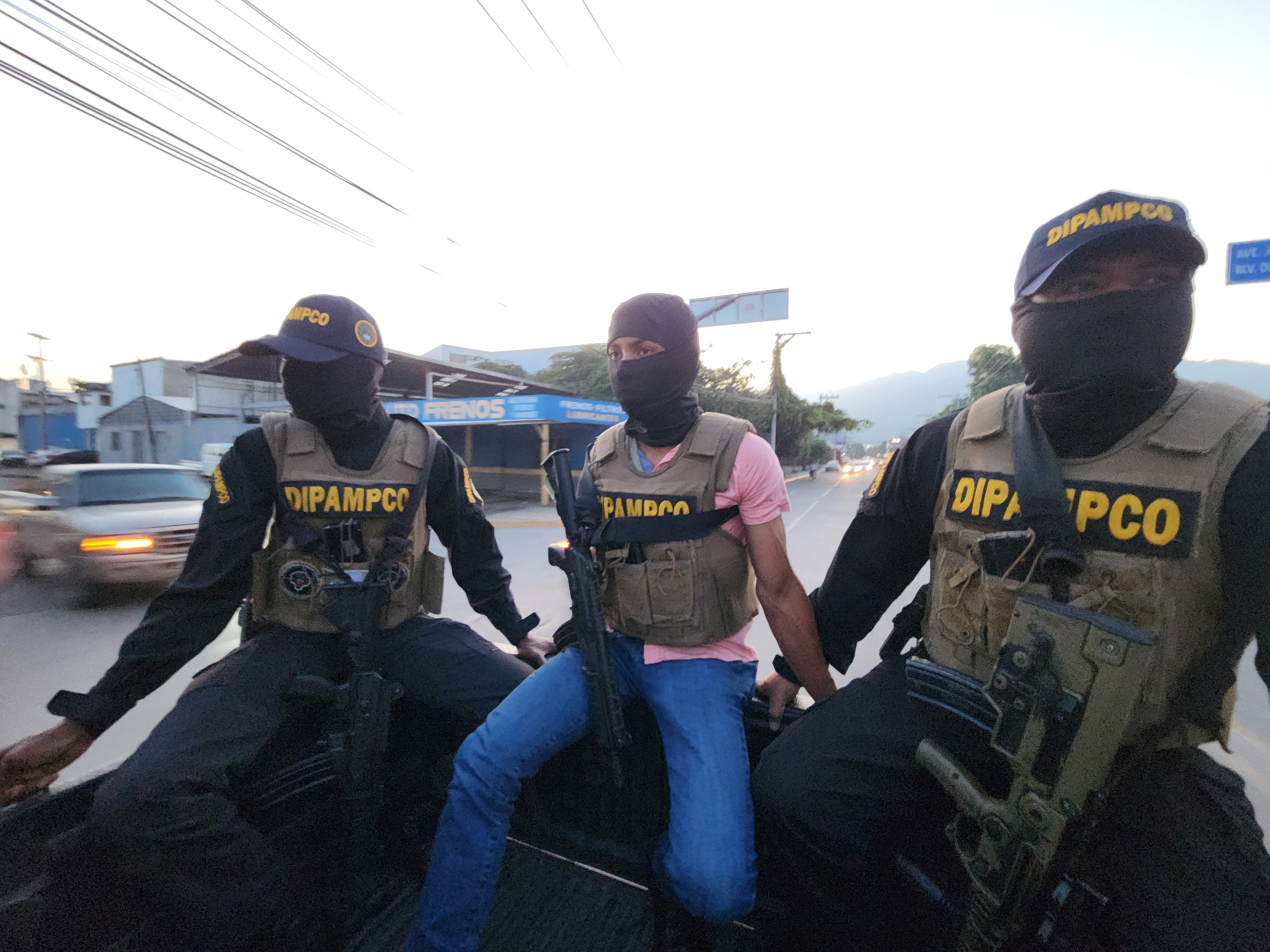 Armed DIPAMPCO members in helmets, face-concealing masks and bulletproof vests ride in the back of an open truck during the day.