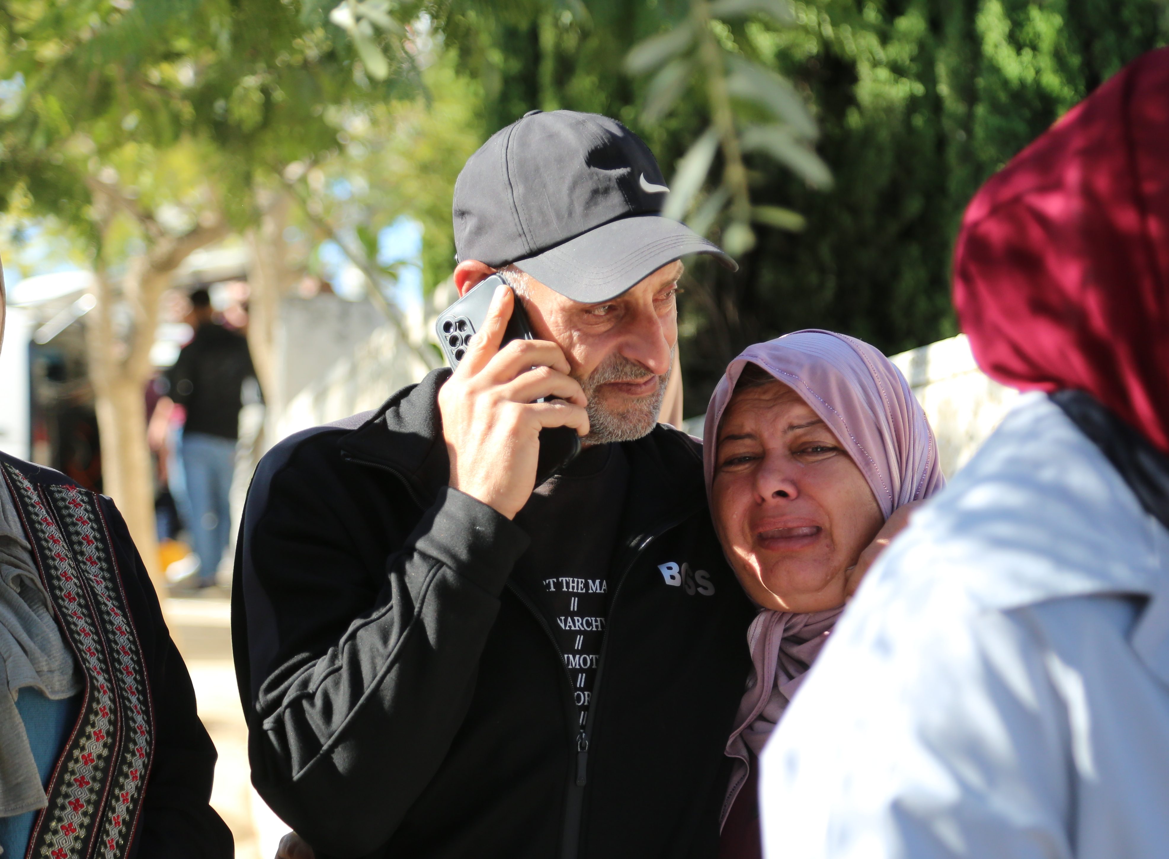 Raed wears a baseball cap. he's middle-aged with frey stubble. he's on his phone and has an arm around Ibtisam who is crying