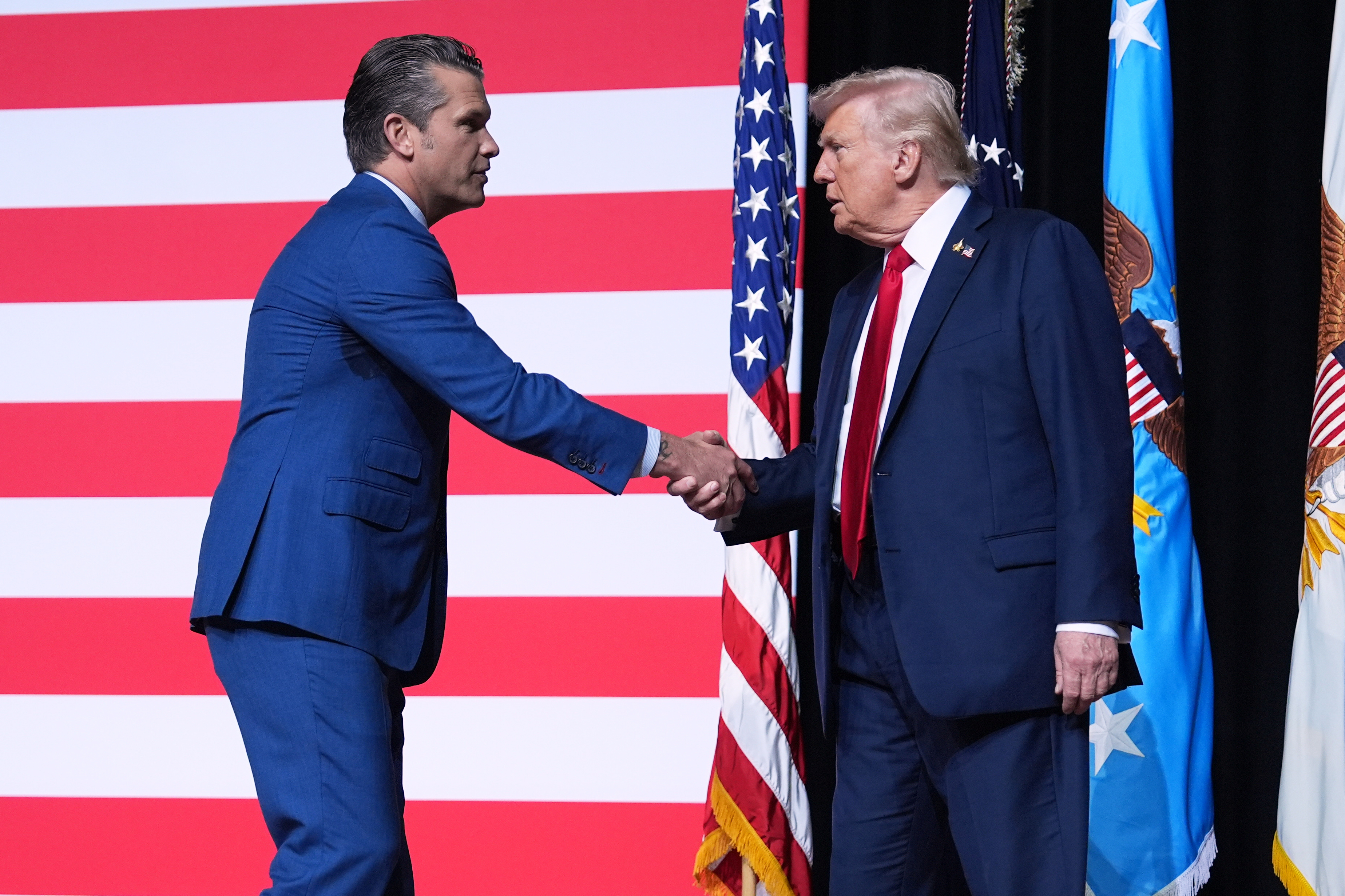 President Donald Trump is greeted by Secretary of Defense Pete Hegseth in front of a US flag.
