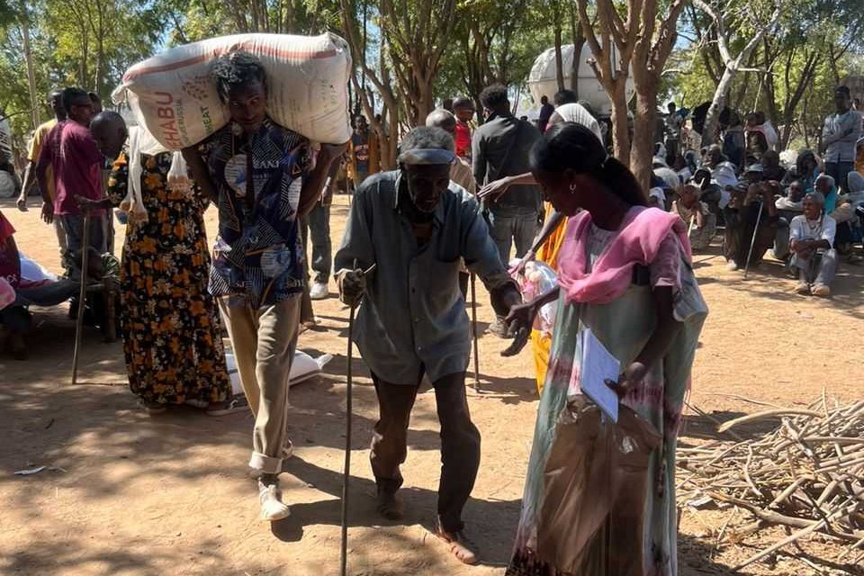 People walk with their belongings, helping an elderly man along with them.