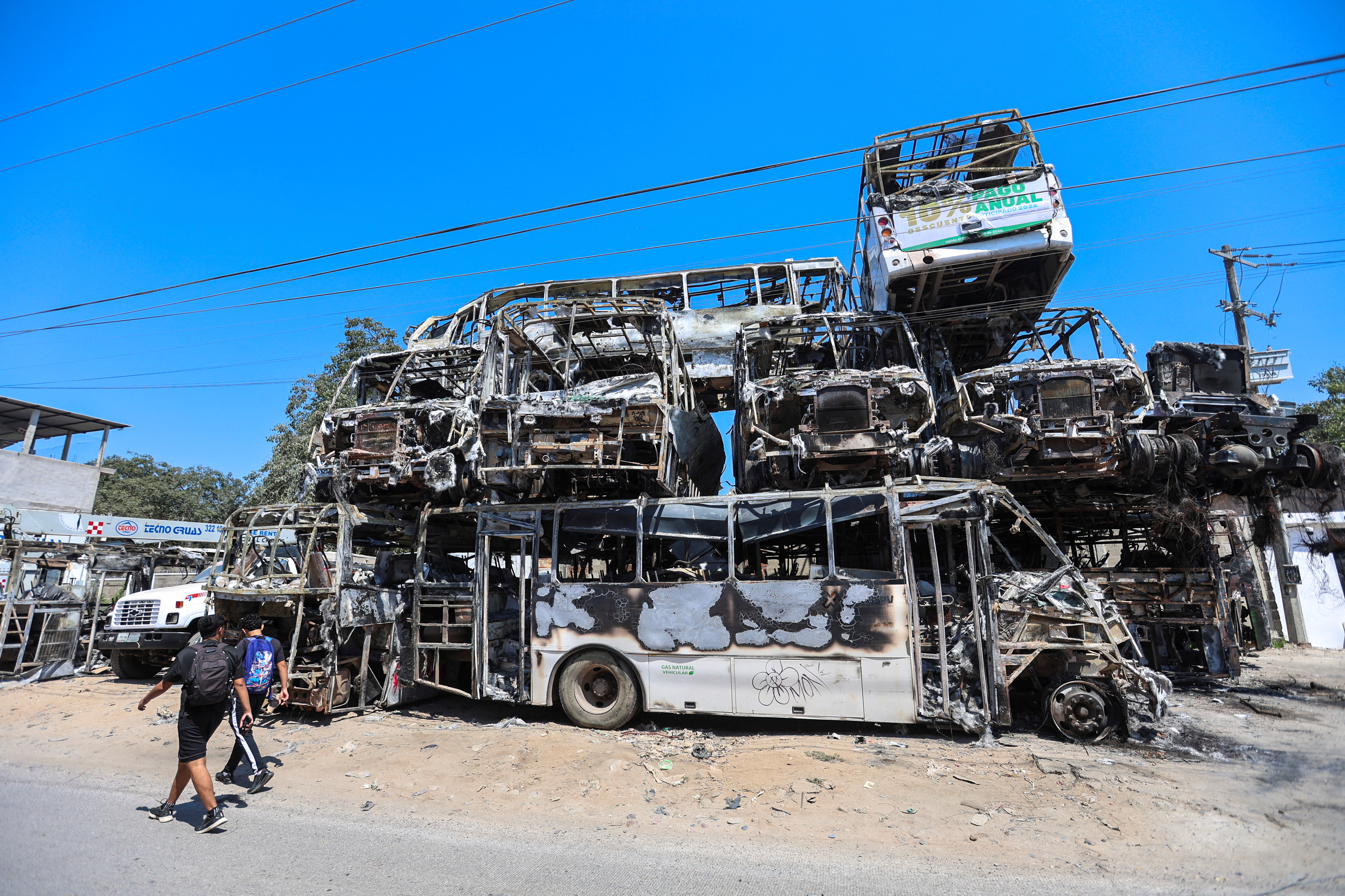Men walk past the charred wreckage of vehicles used in a series of blockades by organized crime following a military operation in which cartel boss Nemesio Oseguera Cervantes, “El Mencho,” was killed on February 22 in the state of Jalisco, at a crane company yard in Puerto Vallarta, Mexico