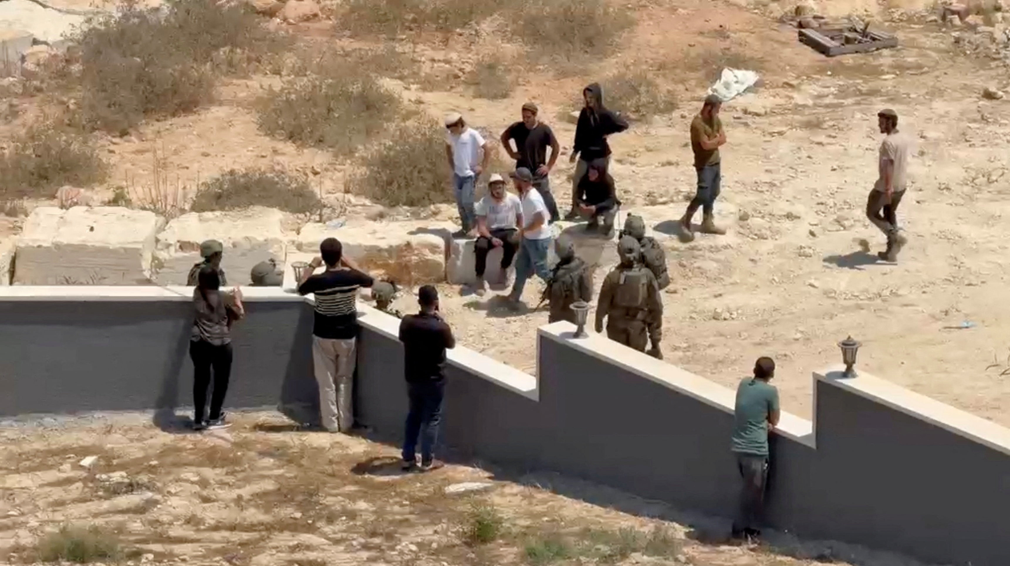 Palestinians use their phones as Israeli settlers and military personnel stand outside a Palestinian home in Qusra, in the Israeli-occupied West Bank