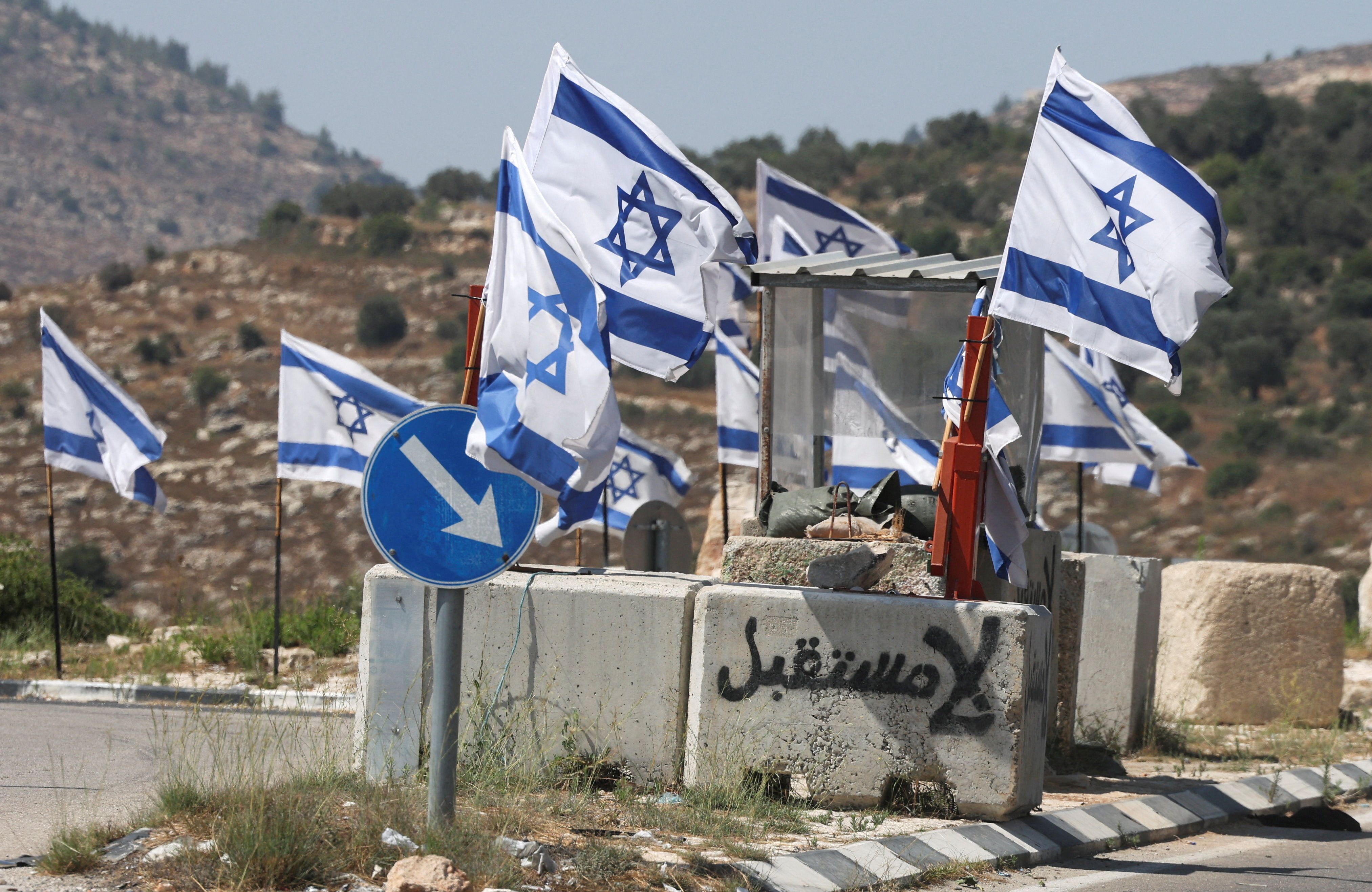 A cement block with the words "no future" spraypainted on it in Arabic, at an Israeli military checkpoint with Israeli flags flying on it.