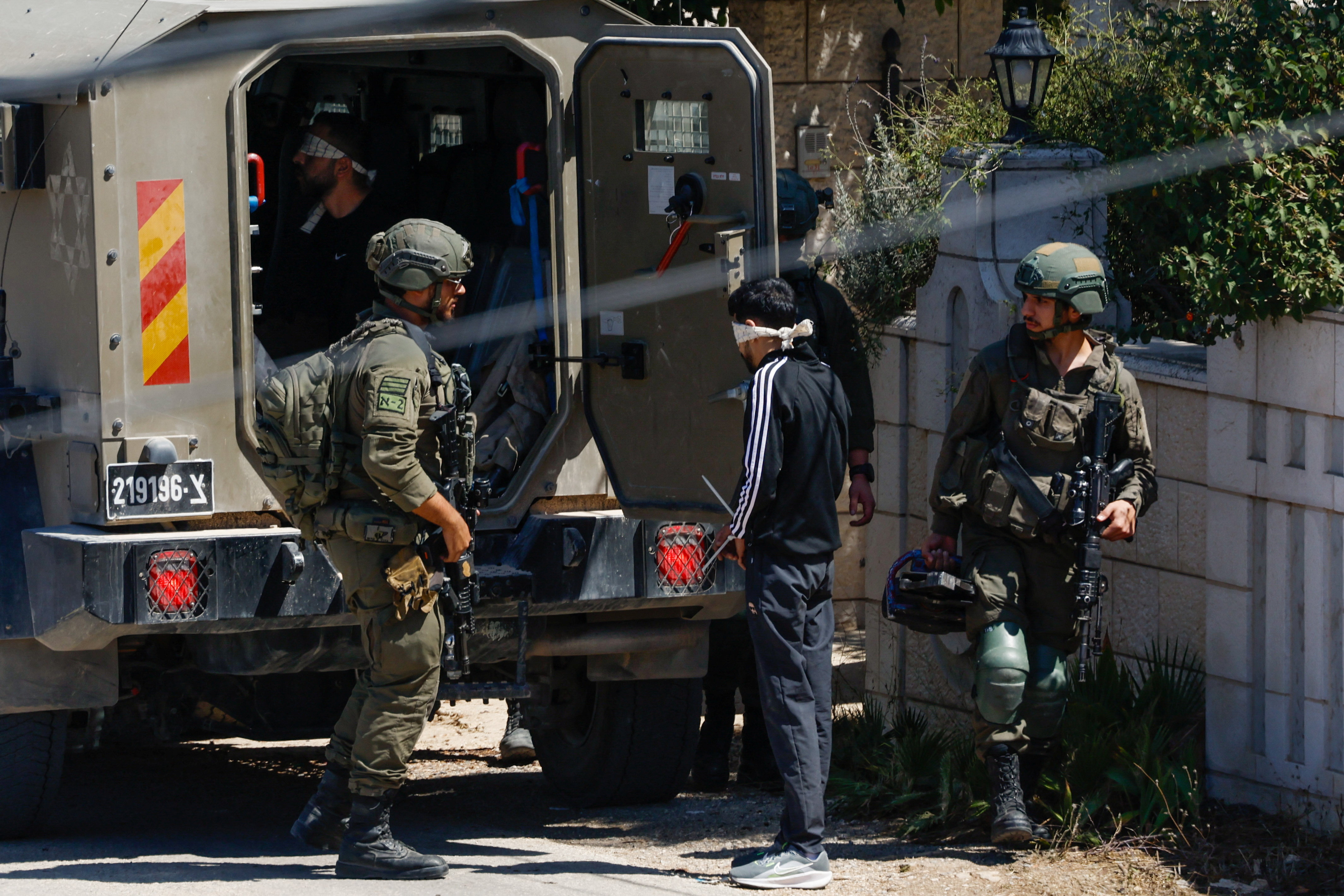 Two soldiers stand with a blindfolded man near a military vehicle.