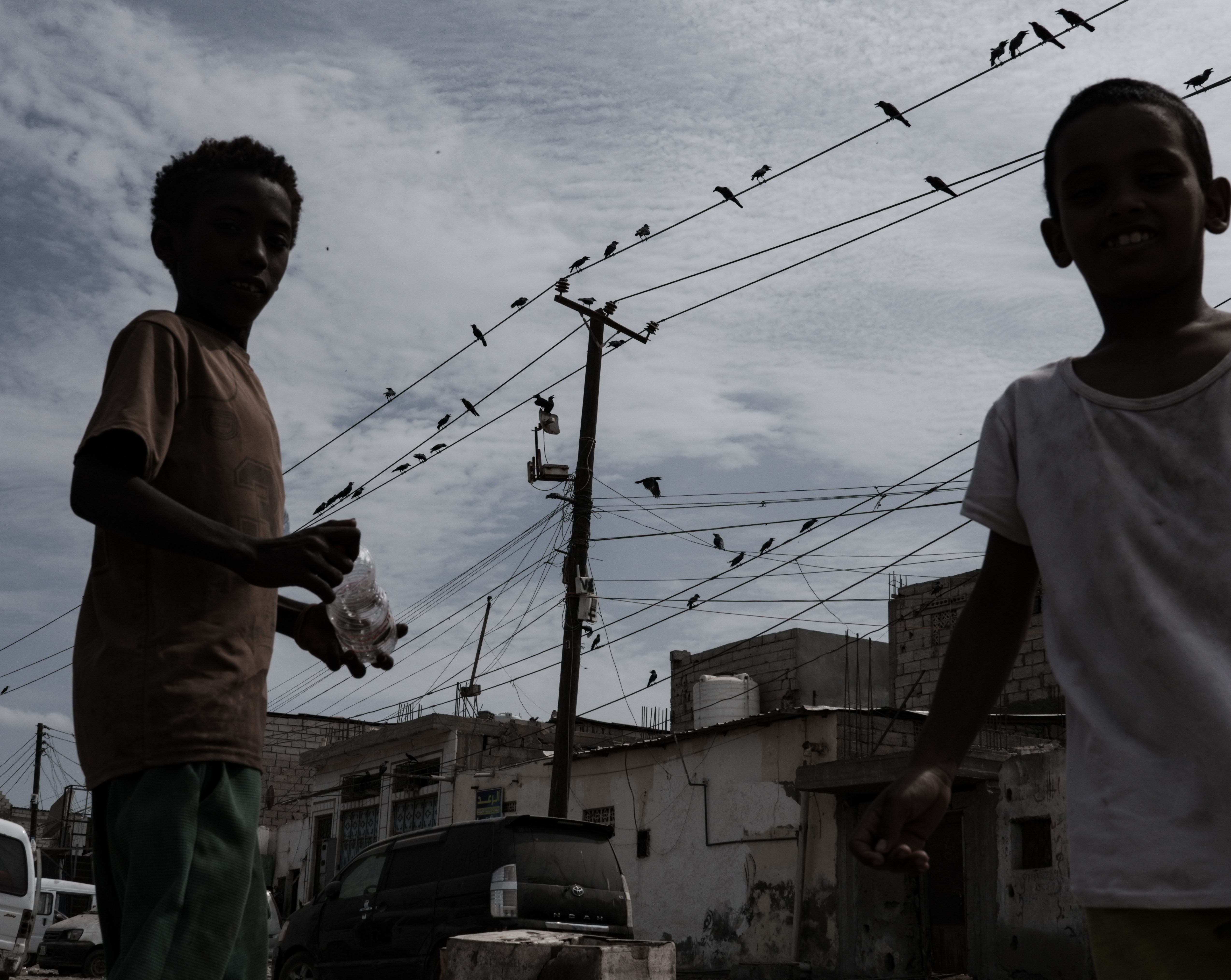 Two children stand beneath electricity lines in a neighborhood in Aden. During long outages, the heat inside homes pushes many children to spend more time outdoors, while families wait for power to return so they can use fans and sleep.