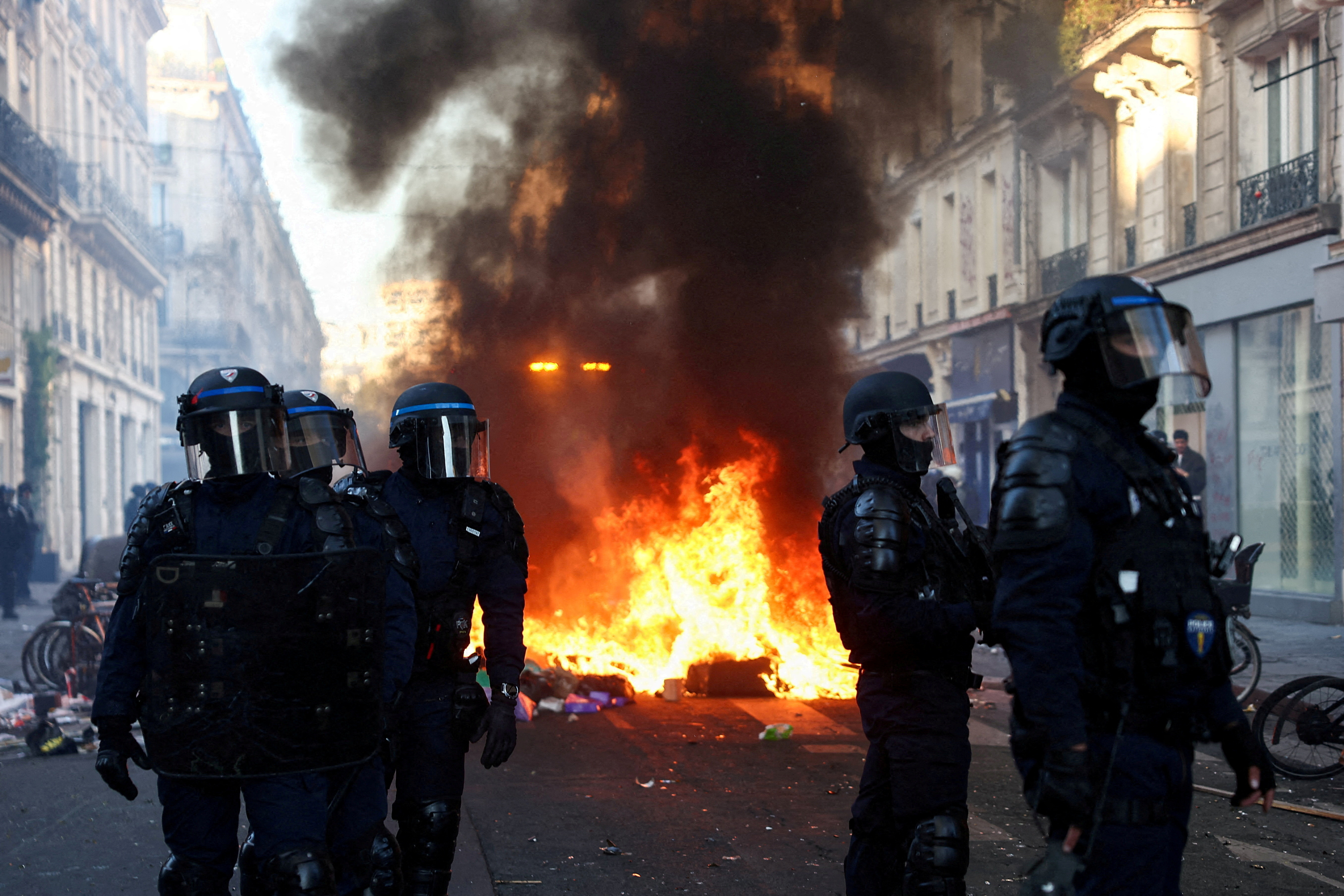 A fire burns as police officers in riot gear stand guard.